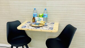 A small dining area with two black chairs, a wooden table, and a floral tablecloth. There are two bottles of water, a bottle of milk, a jar of jam, and a plate with a cake on the table.