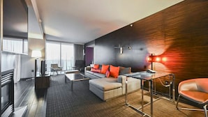 Une chambre d’hôtel moderne, équipée d’un canapé, d’un fauteuil et d’une table basse en verre. On y trouve également un bureau avec une chaise et une lampe. La chambre dispose d’une grande fenêtre et offre une vue sur la ville.