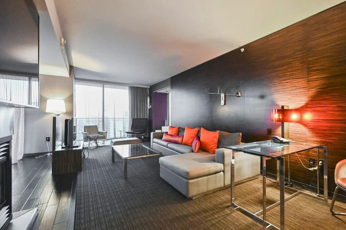 Een moderne hotelkamer met een bank, fauteuil en een glazen salontafel. Er is een bureau met een stoel en een lamp. De kamer heeft een groot raam en uitzicht op de stad.