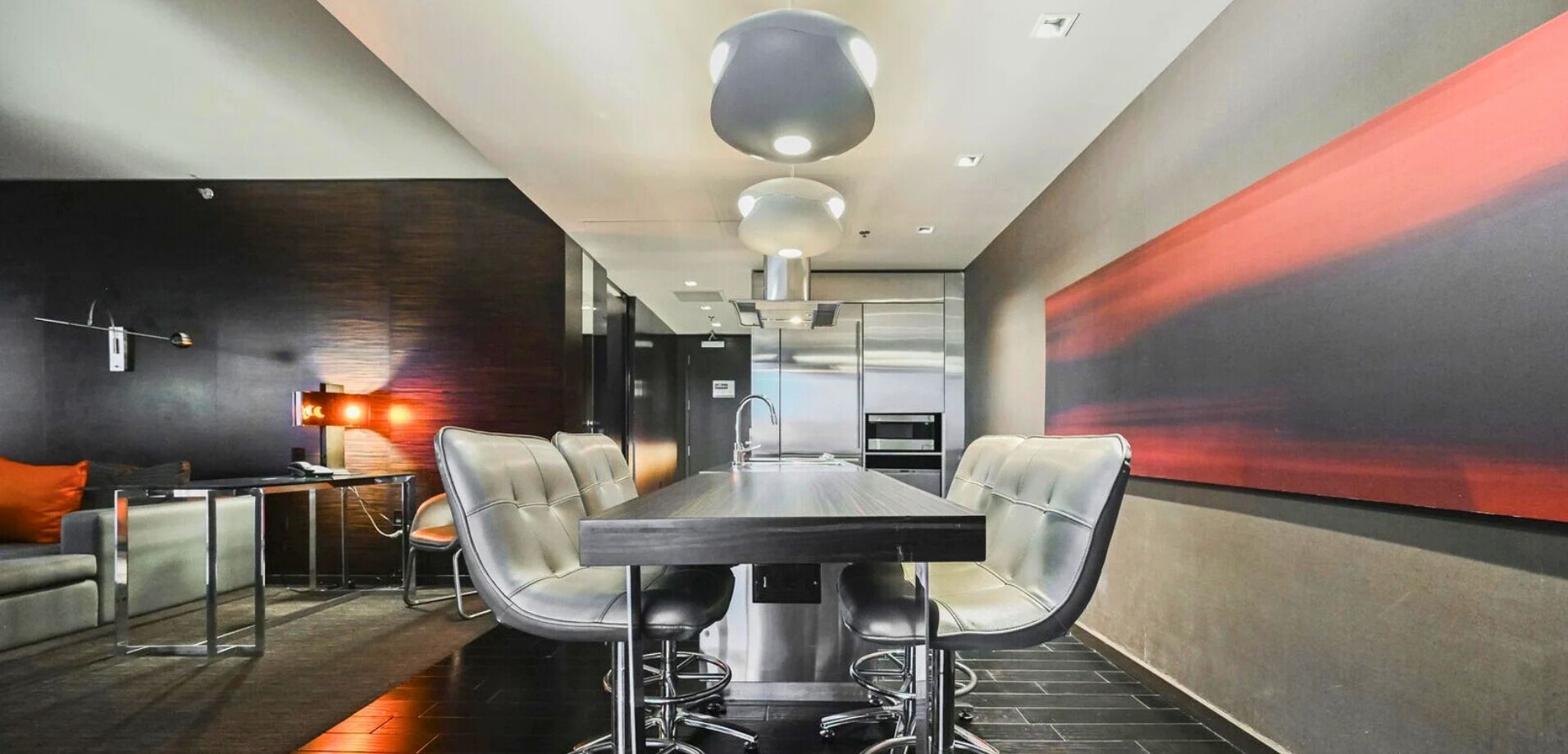 A modern dining area with a long table, four chairs, a bar area with stools, and a large abstract painting on the wall.