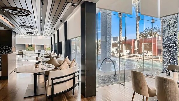 Un espace piscine intérieur moderne doté d’un mur en verre, d’un coin salon avec une table ronde et des chaises, et d’un plafond équipé de luminaires linéaires.
