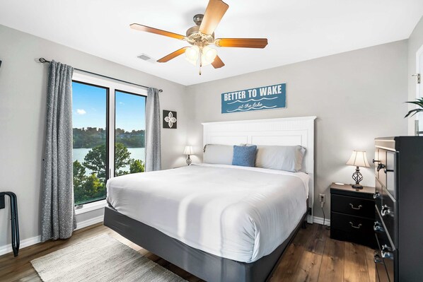 A bedroom with a large bed, a ceiling fan, a view of the lake, and a sign that says "Better to wake at the lake".