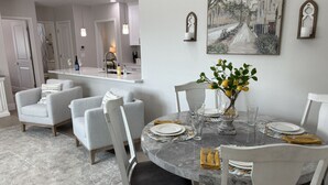 A dining area with a round marble table, white chairs, and a vase with flowers. There is a painting on the wall and a kitchen in the background. - River Trail Stays - Rivers Edge (Tucson)