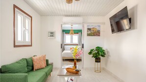 A modern living room with a green sofa, a coffee table with magazines, a large potted plant, and a bed with a green-striped headboard.