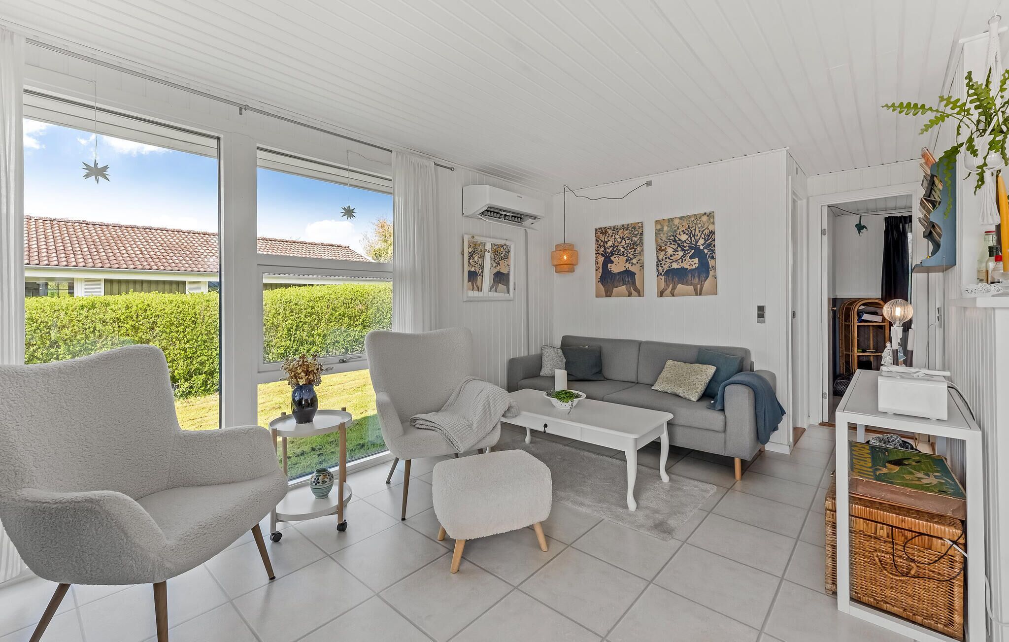 A spacious living room with a grey sofa, a white armchair, and a coffee table. There are large windows providing a view of a garden and a house with a red-tiled roof.