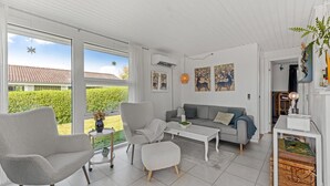 A spacious living room with a grey sofa, a white armchair, and a coffee table. There are large windows providing a view of a garden and a house with a red-tiled roof.