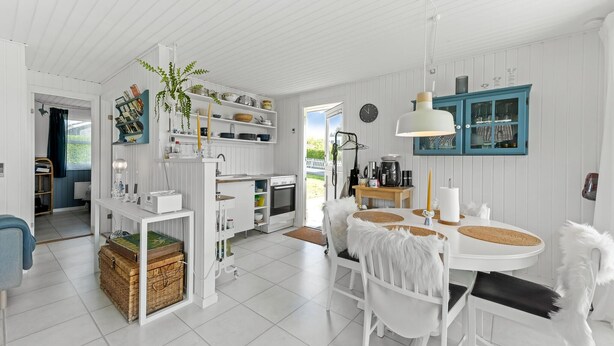 A bright, white interior with a kitchen area featuring white cabinets, a dining table with white chairs, and a hanging light fixture.