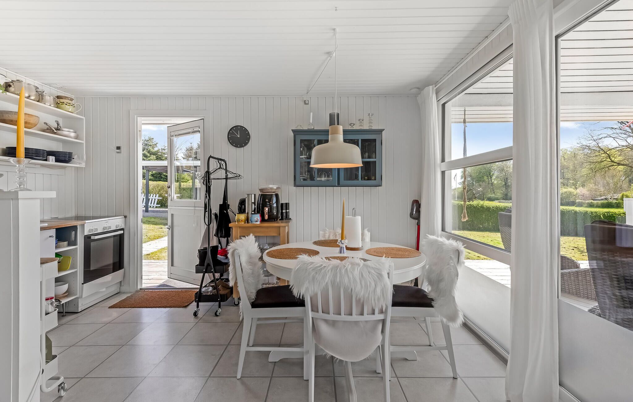 A bright kitchen with white cabinetry, a dining table with white chairs, a hanging light fixture, and a view of the garden through large windows.