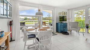 A bright, spacious living area with a large window offering a view of a garden and houses. There is a dining table with chairs, a blue cabinet, and a television.