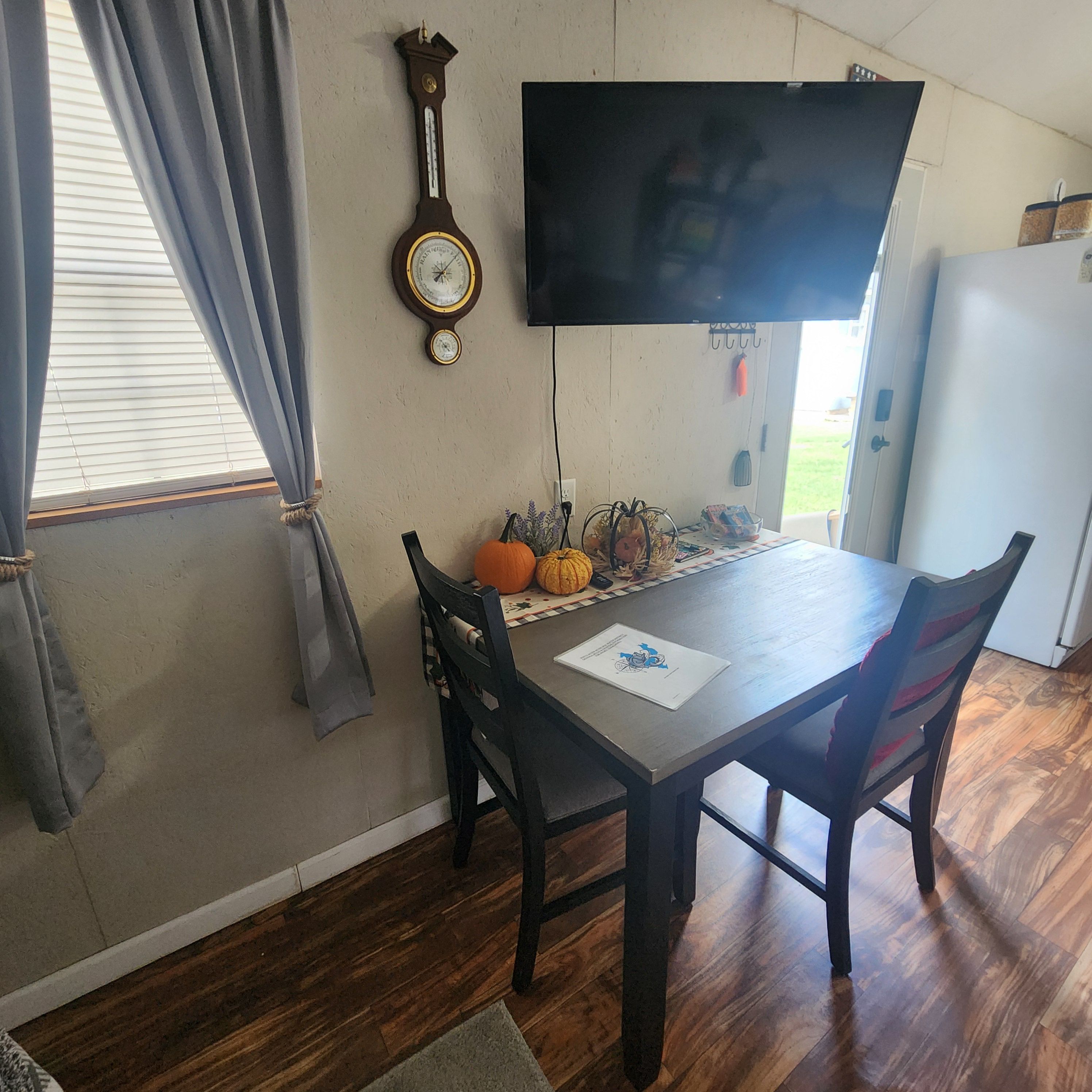A dining area with a wooden table, two chairs, a wall-mounted clock, a television, and a window with curtains.