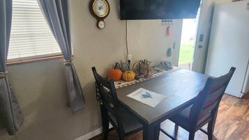 A dining area with a wooden table, two chairs, a wall-mounted clock, a television, and a window with curtains.