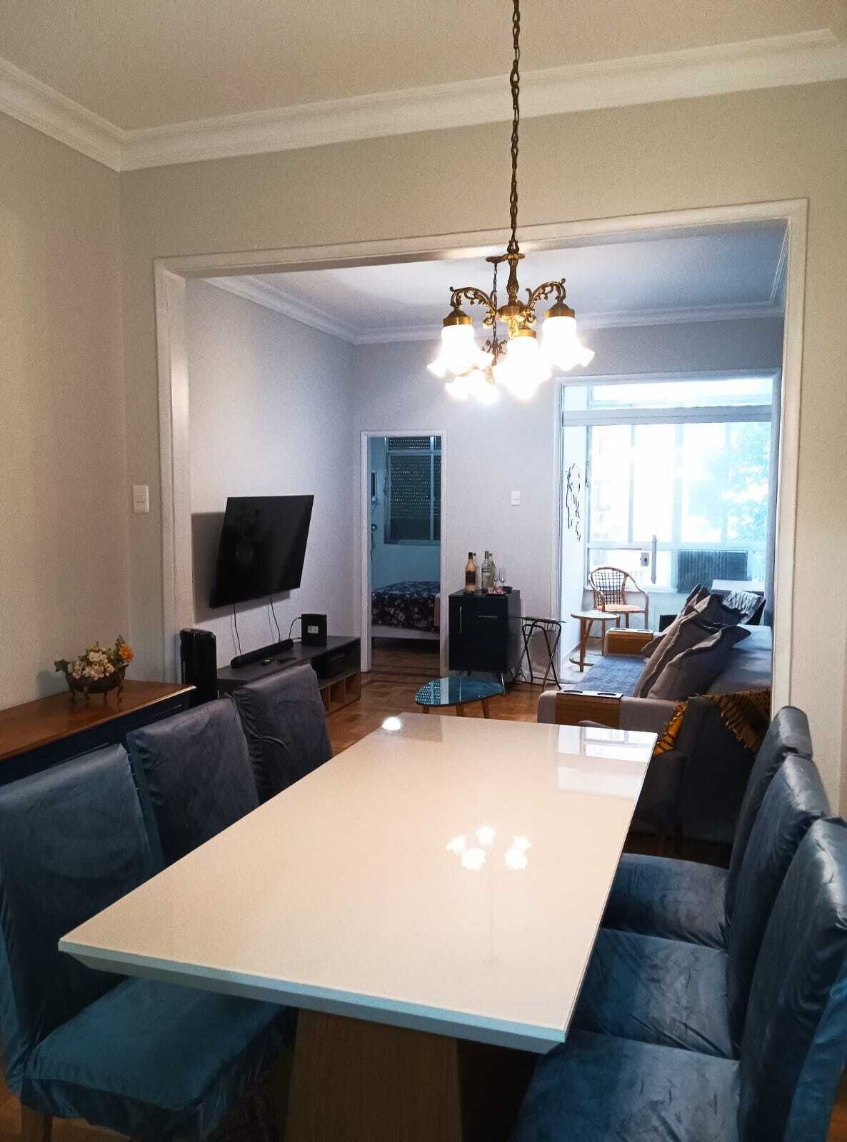 A modern living room with a large dining table, blue leather seating, a TV, and a chandelier.