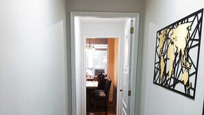 Un pasillo estrecho con piso de madera que conduce a un comedor. A la derecha, una mesa de vidrio con flores, y a la izquierda, una silla de metal negro.