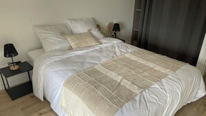 A neatly made bed with a white duvet and a beige quilted throw, a black nightstand with a lamp, and a dark curtain partially drawn to one side. - Country house 20 minutes from Puy du Fou (Mortagne-sur-Sèvre)