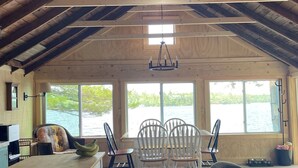 A spacious room with a high ceiling, wooden beams, a chandelier, a dining table with chairs, a large window overlooking a body of water, and a modern chair.