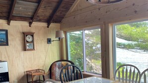 A wooden dining area with a table and chairs, a clock on the wall, and a view of greenery outside.