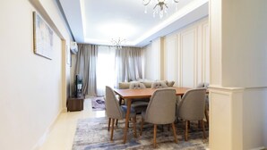 A dining area with a wooden table and six chairs, a chandelier, and a large window with curtains. - 2BRs CityStar Mall Apartment (Al Golf, Nasr City)