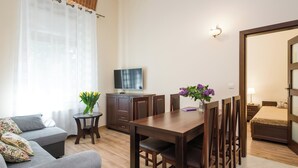 A modern living room with a grey sofa, wooden dining table, and chairs. There is a TV on a cabinet, a small side table with a vase of flowers, and a bed with a wooden frame. - Vanilla 6, 2bdr 2 bth apartment 5min from Krakow's Main Square (Kraków)
