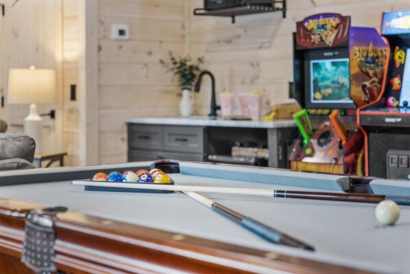 A pool table with balls and a cue stick, a lamp, a potted plant, and arcade games in the background.