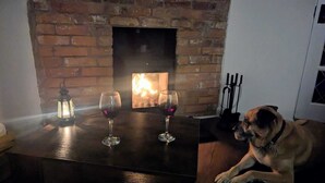 Um quarto com lareira de tijolos, dois copos de vinho tinto sobre uma mesa, um lampião aceso e um cão sentado perto da lareira.