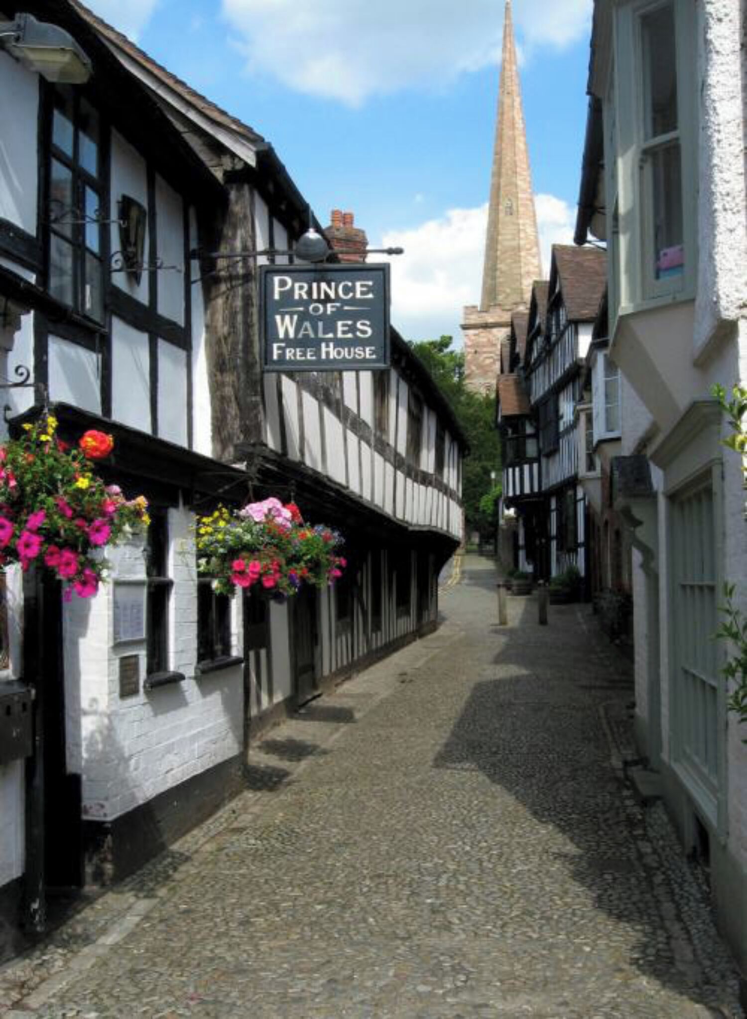 A narrow cobblestone street lined with traditional half-timbered buildings, including a sign for 'Prince of Wales Free House'.