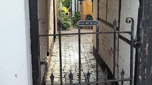Uma estreita viela de calçada que leva a um edifício com portão de ferro preto e uma placa com a inscrição 'Feche o portão'.