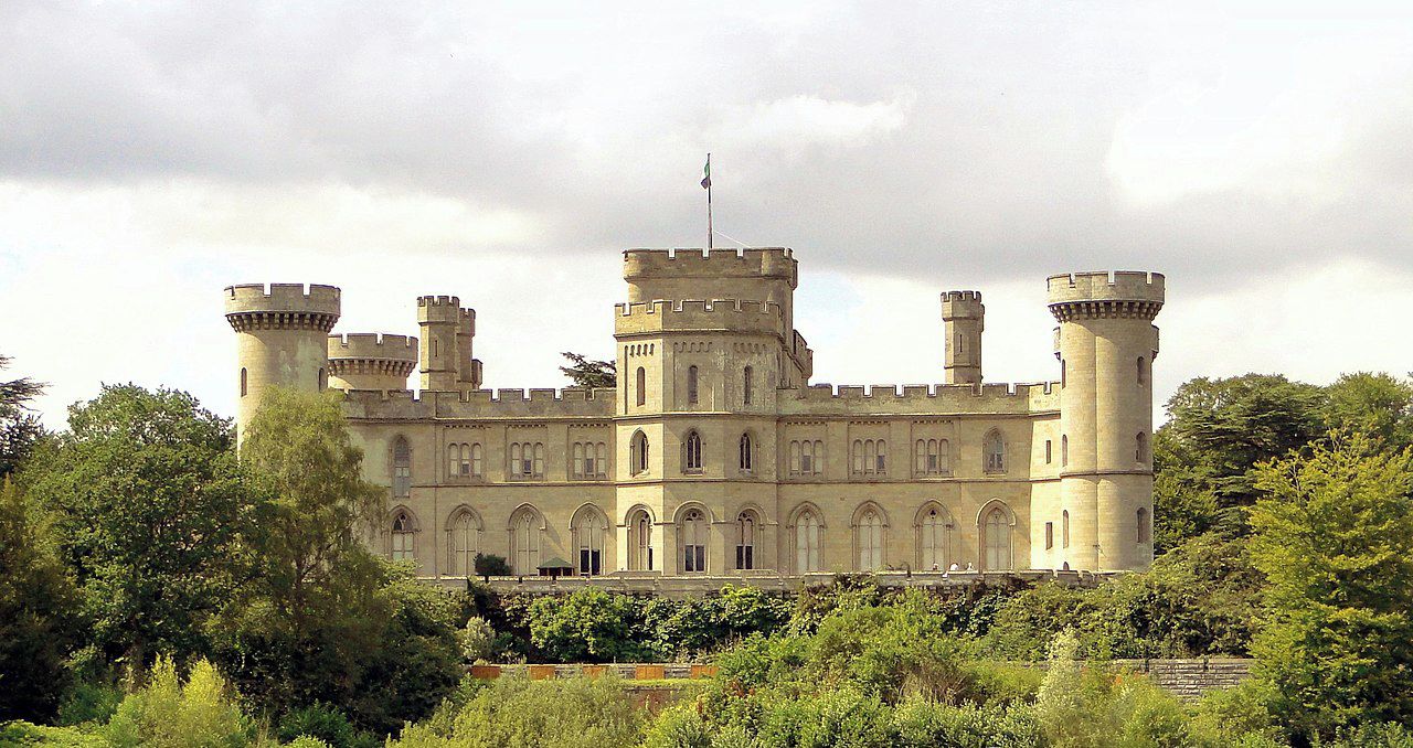 Eastnor Castle with a prominent central tower, surrounded by lush greenery and a body of water in the foreground.