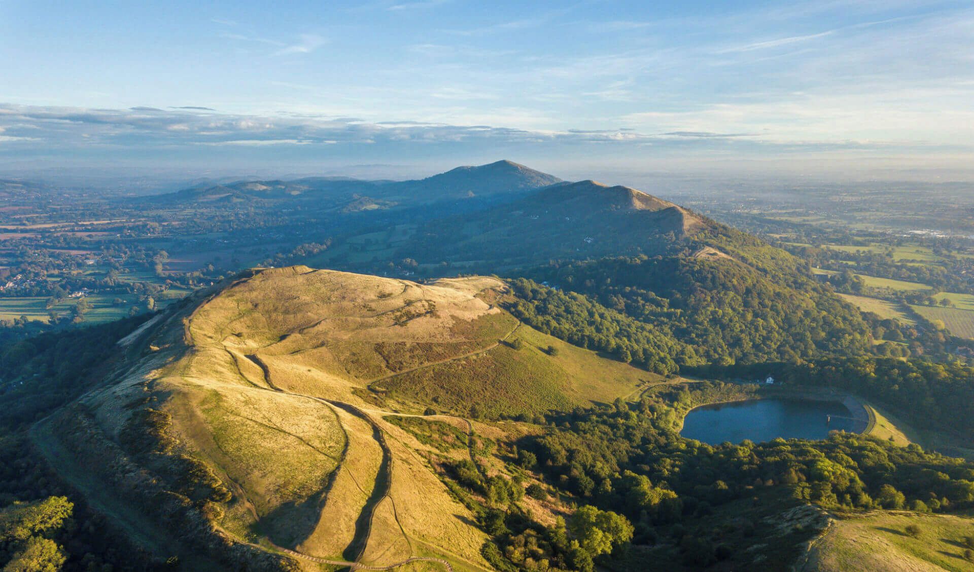 Luftaufnahme der Malvern Hills mit gewundenen Pfaden und einem See.