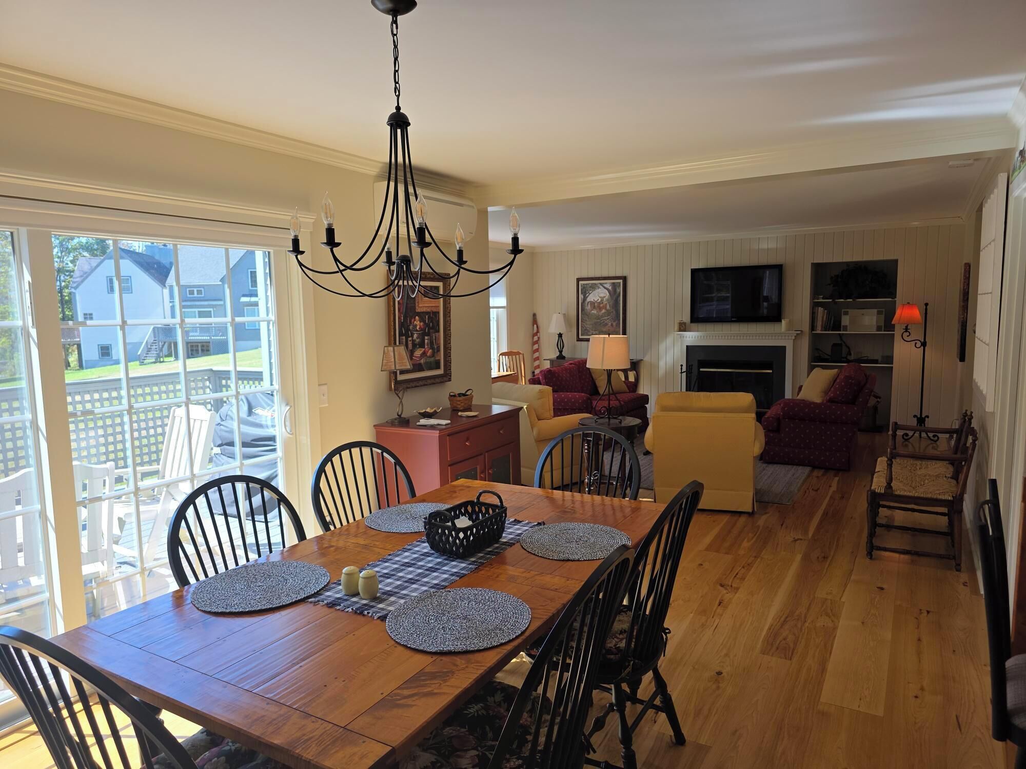 A dining area with a wooden table, black chairs, and a chandelier. There is a living room with a fireplace, sofas, and a TV.