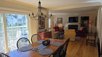 A dining area with a wooden table, black chairs, and a chandelier. There is a living room with a fireplace, sofas, and a TV.
