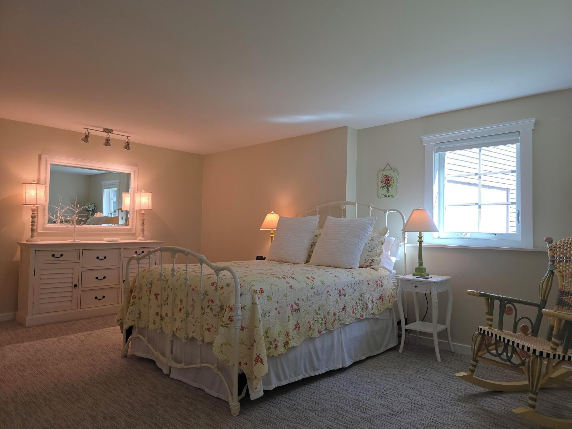 Une chambre à coucher comprenant un lit, des tables de chevet, une commode, un fauteuil à bascule et une fenêtre avec des stores.