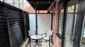 Een klein terras buiten met twee metalen stoelen en een ronde tafel, omringd door een bakstenen muur en een zwart metalen hek.