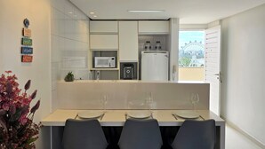 A modern kitchen with white cabinets, a microwave, and a refrigerator. There are three chairs and a table with wine glasses.