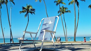 A beach chair with a striped backrest and metal frame, positioned on a concrete platform with palm trees and the ocean in the background.