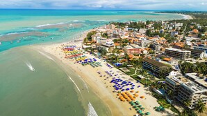 Vista aérea de un complejo turístico con una extensa playa de arena, numerosas sombrillas y diversos edificios, incluyendo hoteles y residencias.