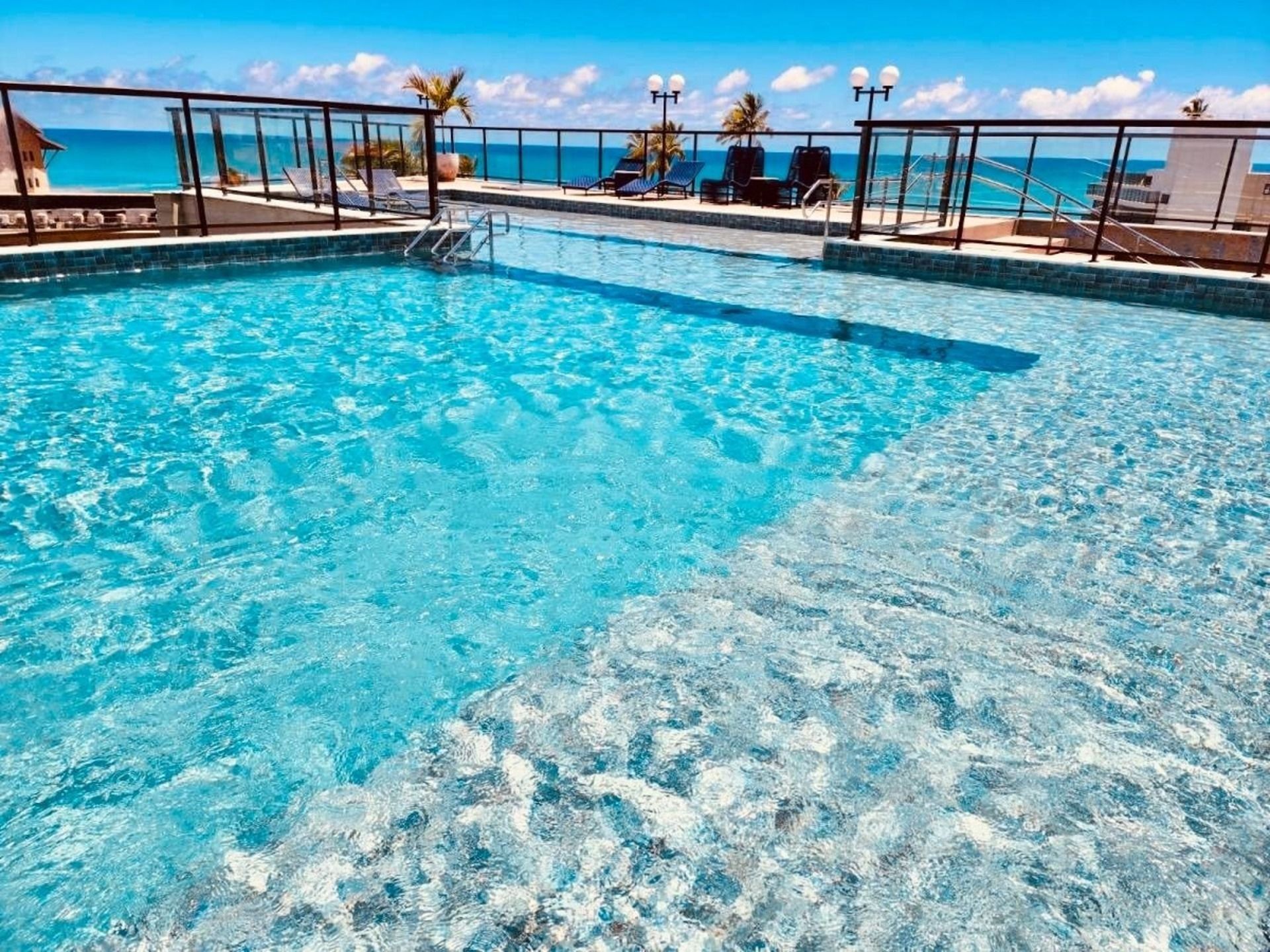 Piscina azul e límpida com vista para o oceano, rodeada por espreguiçadeiras e um guarda-corpo de vidro.