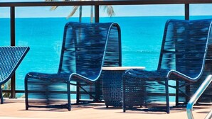 Un balcón con dos sillas de mimbre azules y una mesita pequeña, con vista a un mar azul y despejado donde se distingue una palmera al fondo.