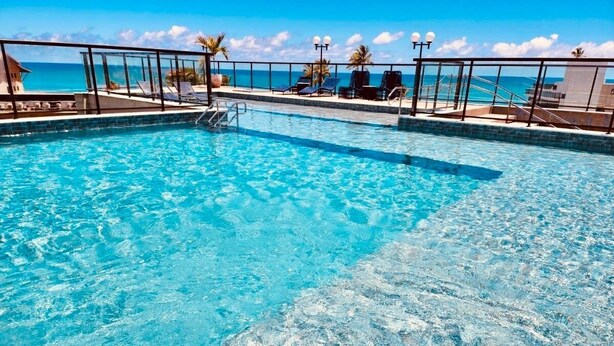 Una piscina azul cristalina con una barrera de vidrio, sillas de descanso y vistas al océano.