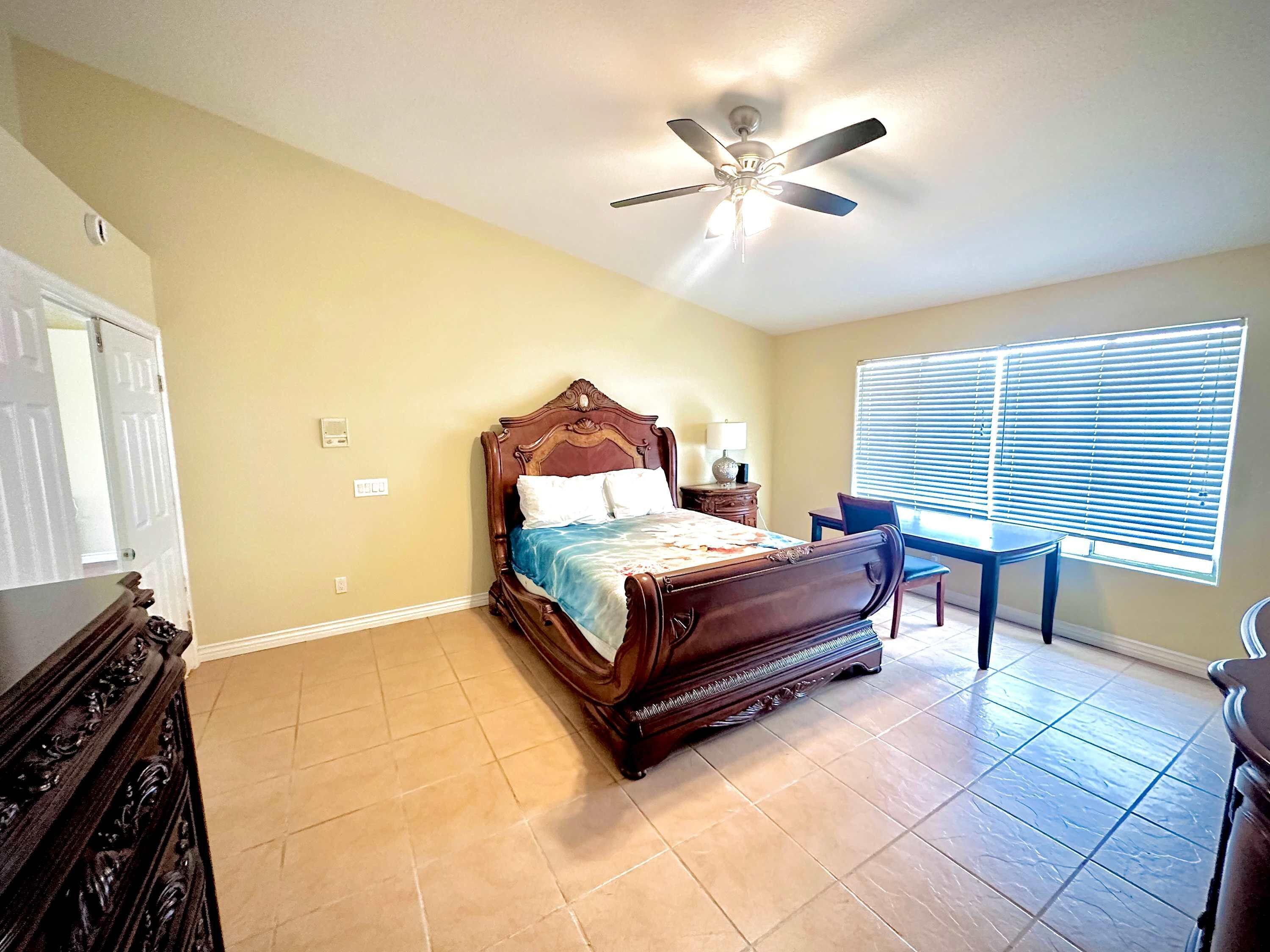 A bedroom with a wooden bed, a ceiling fan, a window with blinds, and a small table with a chair.