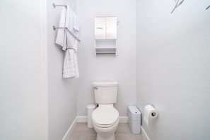 A modern bathroom with a white toilet, a towel rack with folded towels, a small mirror, and a trash bin.