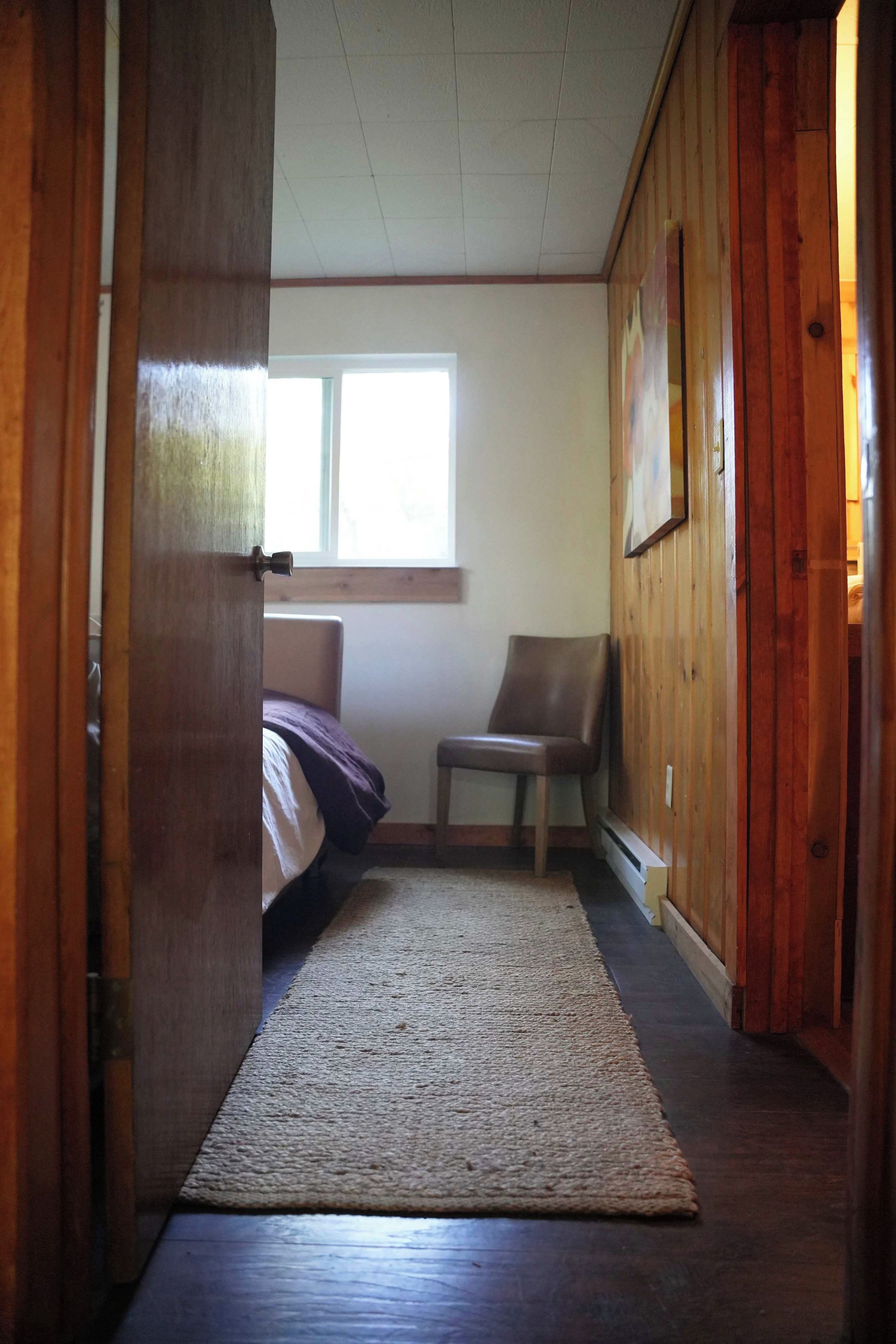 A narrow hallway with a wooden door on the left, a bed with a purple blanket, a chair, and a window providing natural light.