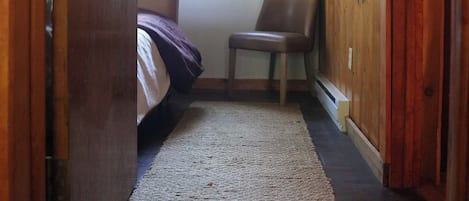 A narrow hallway with a wooden door on the left, a bed with a purple blanket, a chair, and a window providing natural light.