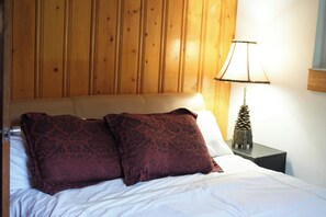 Ein Bett mit zwei dunkel gemusterten Kissen, einem Holzkopfbrett, einer weißen Bettdecke und einem Nachttisch mit einer Lampe und einer Kiefernzapfen-Dekoration.