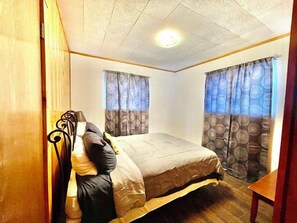 A small, cozy bedroom with a single bed, two windows with patterned curtains, and a wooden headboard.