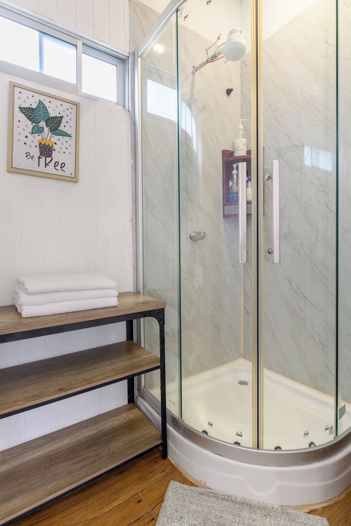 A modern bathroom with a glass-enclosed shower, a wooden bench, and a potted plant with the text "Be Free".