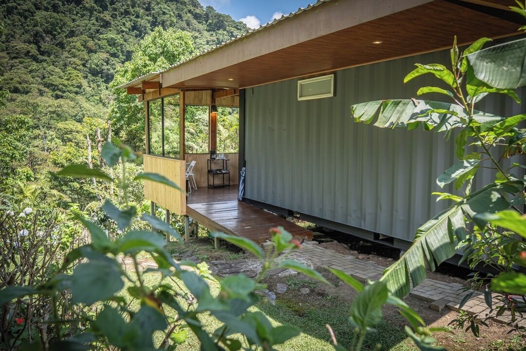 A modern cabin with a wooden deck and large windows, surrounded by lush greenery.