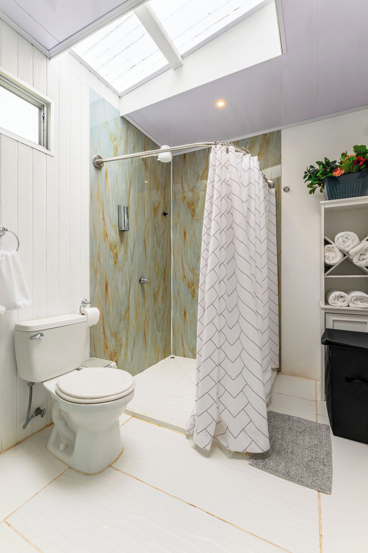 A modern bathroom with a skylight, a shower with a chevron-patterned curtain, a toilet, and a shelf with towels.