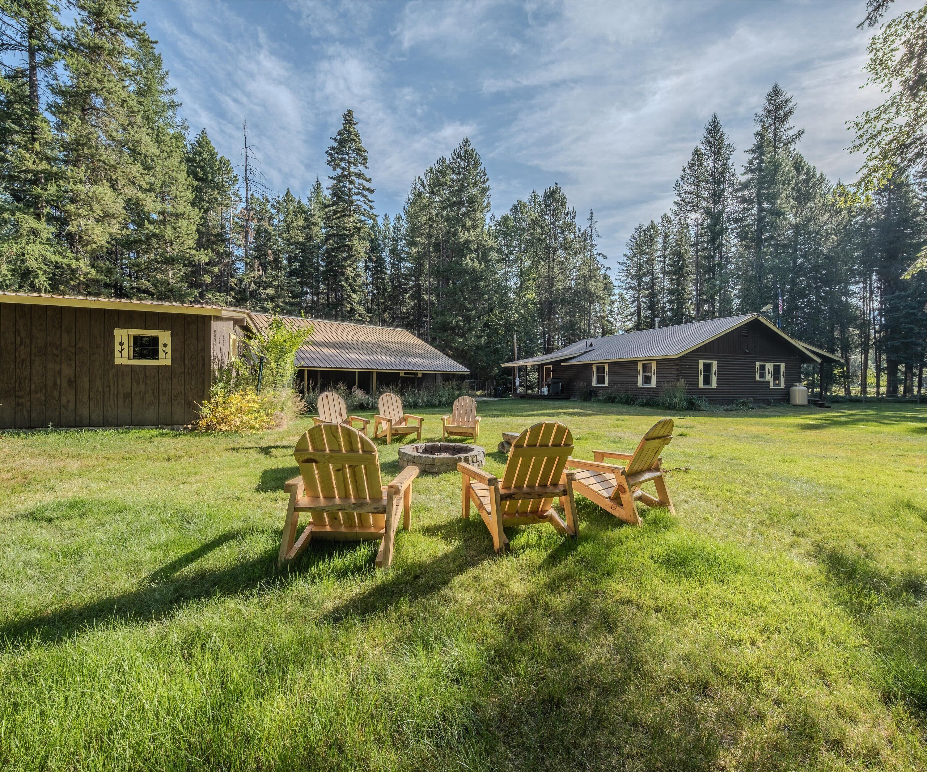 A rustic cabin with a metal roof, surrounded by a grassy yard with Adirondack chairs and a fire pit.