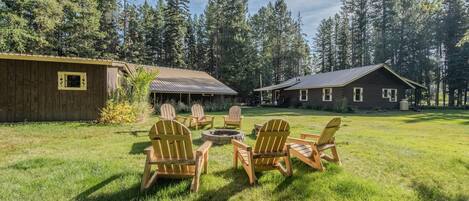 A rustic cabin with a metal roof, surrounded by a grassy yard with Adirondack chairs and a fire pit.