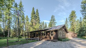 Une cabane rustique en rondins de bois, dotée d’un porche, entourée d’une allée gravillonnée et d’une zone herbeuse.